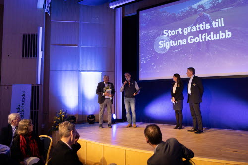 Fyra personer står på scen - två delar ut ett pris till de övriga två. I bakgrunden syns en bild med texten Stort grattis till Sigtuna Golfklubb.